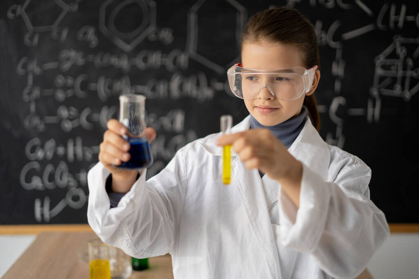 маленька вчена дівчинка в окулярах в лабораторному пальто з хімічними колбами, на шкільному фоні з науковими формулами, назад до концепції школи
 - Фото, зображення