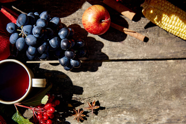 秋の背景:木のテーブルの上に高い角度の影を持つブドウ、リンゴ、ビブラム、スグリ、紅茶とシナモンスティックのマグカップ。感謝祭のコンセプト。屋外の秋の審美的な。スペースのコピー. - 写真・画像