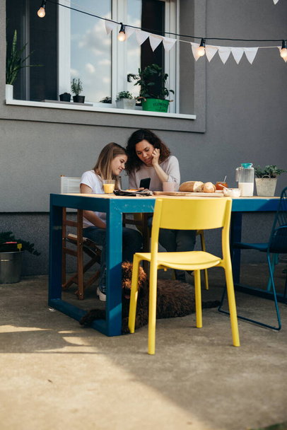 mother and daughter eating breakfast and talking in their home backyard. they are using smartphone - Foto, imagen