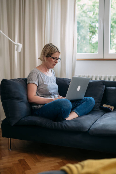 middle aged caucasian woman using laptop computer sits on sofa in living room - Photo, Image