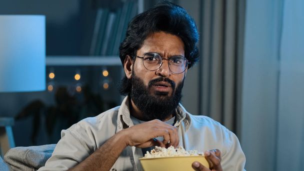 Interested attentive Arabic Hispanic Indian bearded man guy in glasses eating popcorn watching TV show series horror movie late night feeling scary shocked scared reaction disgust horrible thriller - Photo, Image