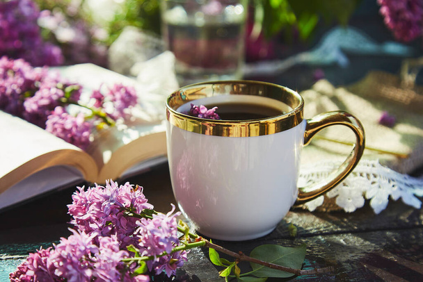 ロマンチックな日当たりの良い庭、コーヒーを飲みながらリラックスしてレジャー。白いカップをモックアップし、ライラックの花に囲まれた本。コーヒー、ライラックの花や本のカップ。ハードシャドウだ。夏の日、審美的な庭. - 写真・画像