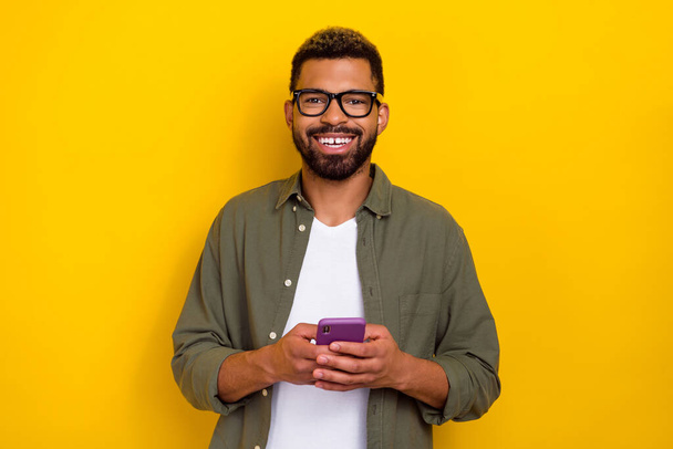 Photo of positive attractive guy dressed green shirt eyewear communicating modern gadget isolated yellow color background - Foto, Imagen