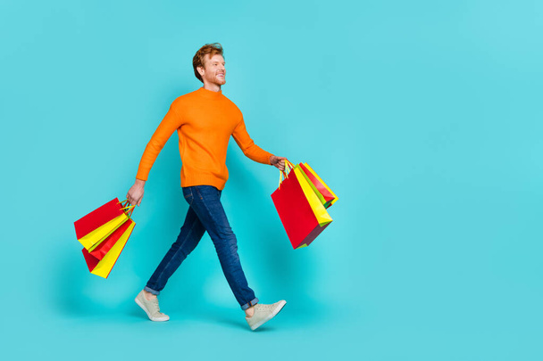 Full length photo of funny dreamy guy dressed orange sweater walking holding bags empty space isolated turquoise color background - Fotoğraf, Görsel