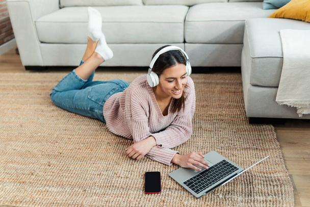 Shot of pretty young woman listening music with headphone while working with laptop lying on the floor at home. - Photo, Image