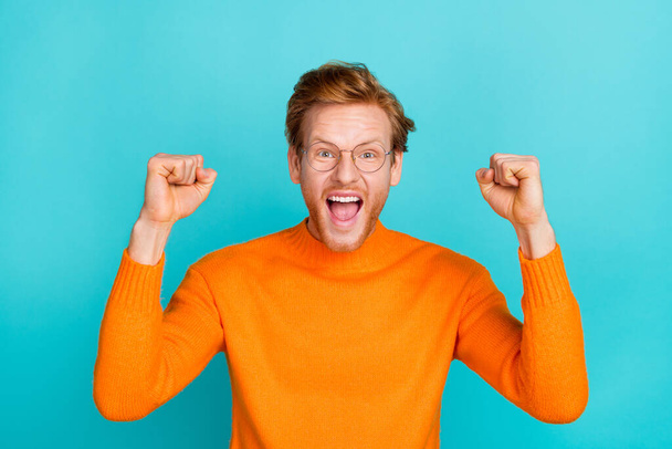 Photo of cute lucky guy dressed orange sweater eyewear screaming rising fists isolated turquoise color background - Фото, изображение