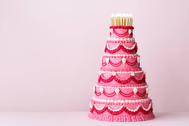Extravagante roze tiered verjaardagstaart versierd met vintage boterroom piped franjes en gouden verjaardagskaarsen - Foto, afbeelding