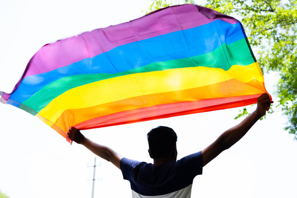 back view shot of young man proudly holding waving lgbtq flag - cocnept of pride month celebration, support and movement. - Photo, Image