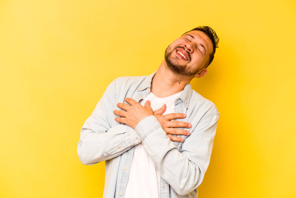 Νεαρός Ισπανός απομονωμένος σε κίτρινο φόντο γελώντας κρατώντας τα χέρια στην καρδιά, την έννοια της ευτυχίας. - Φωτογραφία, εικόνα