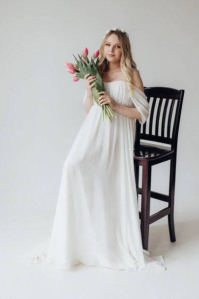 блондинка вагітна жінка в білій сукні в студії на білому тлі, що тримає букет квітів тюльпанів
 - Фото, зображення