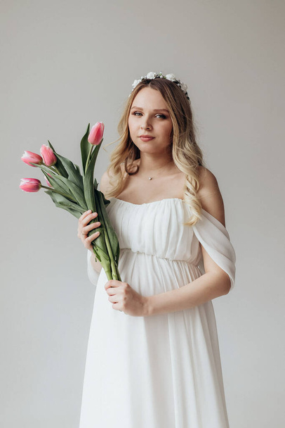 blonde pregnant woman in a white dress in the studio on a white background holding a bouquet of tulip flowers - Foto, immagini