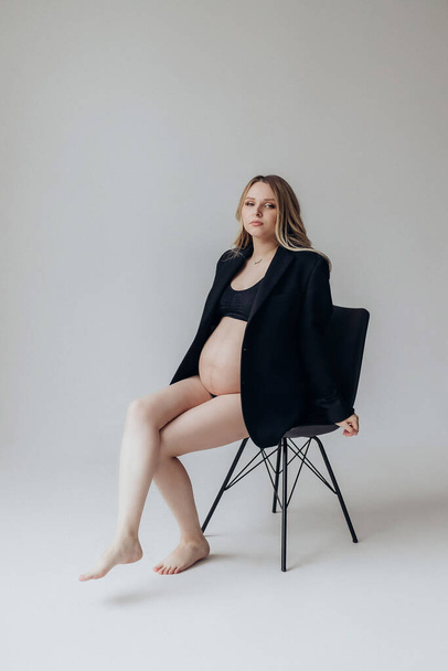 egy szőke terhes nő fekete kabátban és fehérnemű ül egy széken a stúdióban, fehér alapon tartja a kezét a hasán. - Fotó, kép