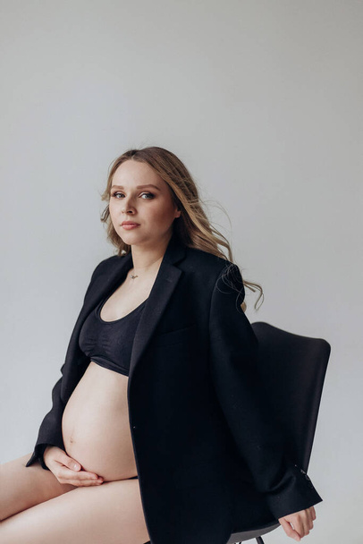 une femme enceinte blonde vêtue d'une veste noire et de la lingerie assise sur une chaise dans le studio sur fond blanc tient sa main sur le ventre - Photo, image