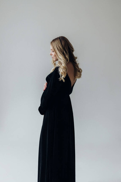 a pregnant blonde woman in a black dress stands in the studio on a white background holds her hand on her stomach - Photo, Image
