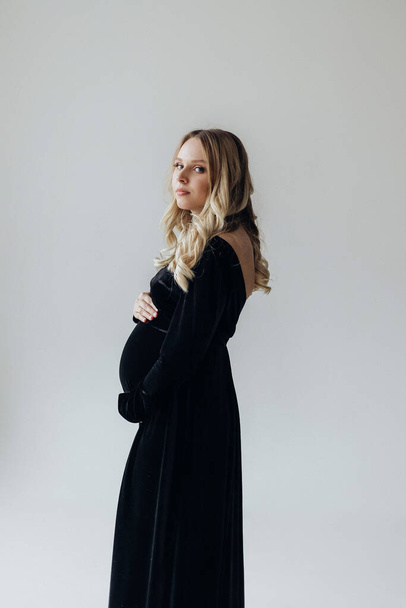 вагітна блондинка в чорній сукні стоїть в студії на білому тлі тримає руку на животі
 - Фото, зображення