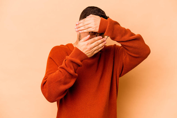 Young hispanic man isolated on beige background blink at the camera through fingers, embarrassed covering face. - Photo, Image