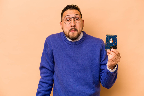 Hispanic informatic man isolated on beige background shrugs shoulders and open eyes confused. - 写真・画像
