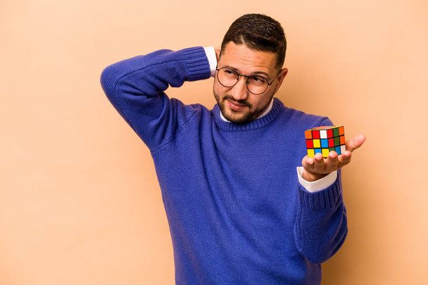Hispanic man holding a Rubik cube isolated on beige background touching back of head, thinking and making a choice. - Fotó, kép