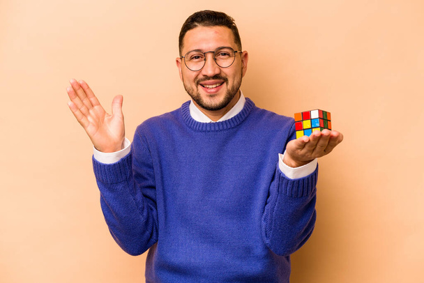 Ισπανόφωνος κρατώντας ένα κύβο Rubik απομονώνονται σε μπεζ φόντο λαμβάνουν μια ευχάριστη έκπληξη, ενθουσιασμένοι και σηκώνοντας τα χέρια. - Φωτογραφία, εικόνα