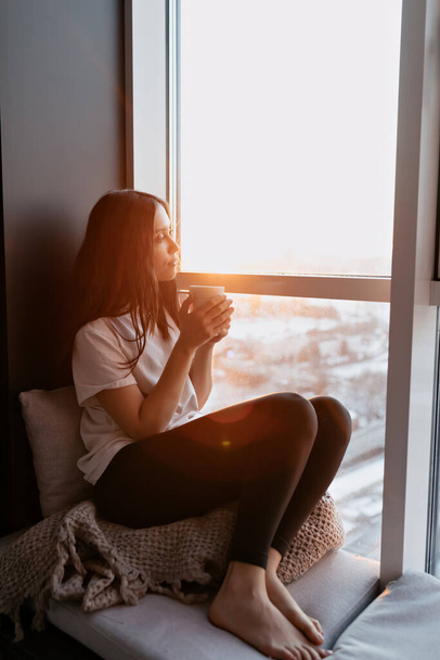 Full-length foto van ontspannen charmante dame met lang golvend haar in wit t-shirt en zwarte legging zitten in de buurt van het raam in het zonlicht bij zonsondergang met uitzicht op de stad. Hoge kwaliteit foto - Foto, afbeelding