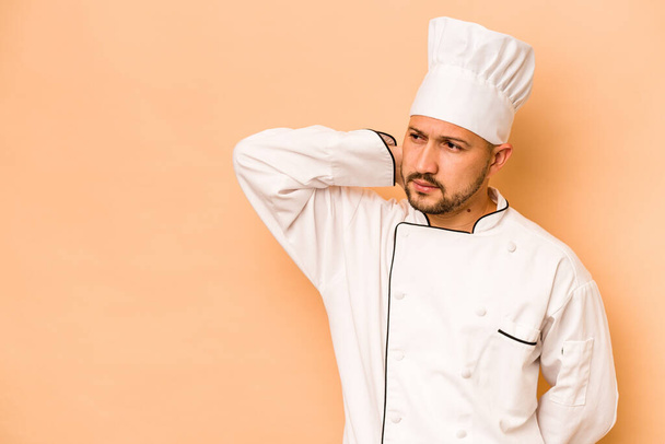 Ισπανόφωνος σεφ άνθρωπος απομονώνονται σε μπεζ φόντο αγγίζοντας πίσω μέρος του κεφαλιού, σκέψης και κάνοντας μια επιλογή. - Φωτογραφία, εικόνα