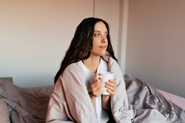 Adorabile bella donna con i capelli scuri ondulati seduta nel letto con una tazza di caffè e guardando da parte alla luce del sole. Carino signora con sorriso felice rilassarsi a casa al mattino  - Foto, immagini