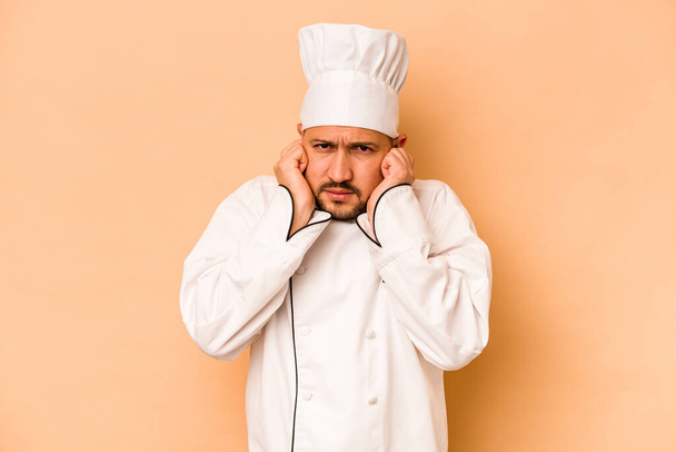 Latynoski kucharz odizolowany na beżowym tle przykrywający uszy dłońmi. - Zdjęcie, obraz
