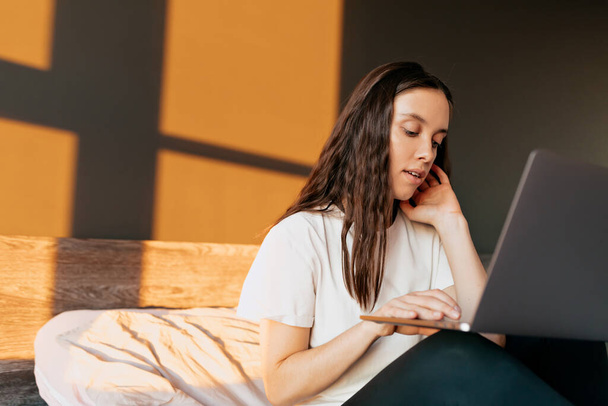 Charmant stijlvol europees meisje met golvend donker haar dragen witte t-shirt kijken naar laptop en werken met een vrolijke glimlach.  - Foto, afbeelding