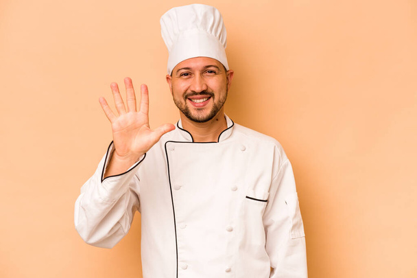 Chef hispanique homme isolé sur fond beige souriant gai montrant numéro cinq avec les doigts. - Photo, image
