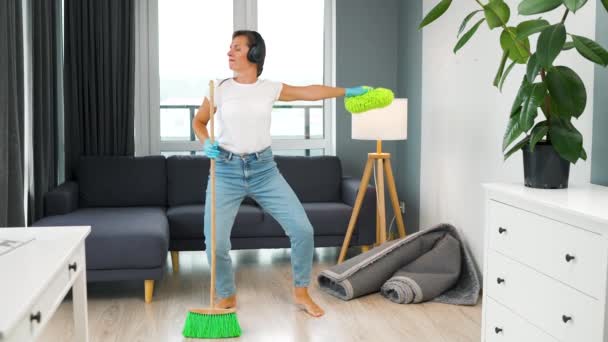 Een vrouw met een koptelefoon die het huis schoonmaakt en plezier heeft met dansen met een bezem en een washandje. Langzame beweging - Video
