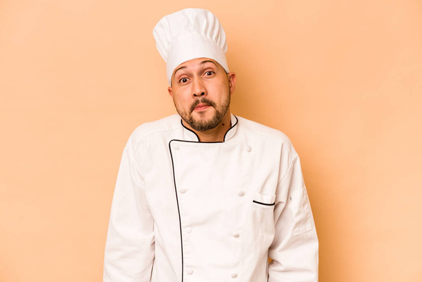 Hispanic chef man isolated on beige background shrugs shoulders and open eyes confused. - Foto, Imagen