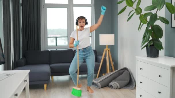 Een vrouw met een koptelefoon die het huis schoonmaakt en plezier heeft met dansen met een bezem. Langzame beweging - Video