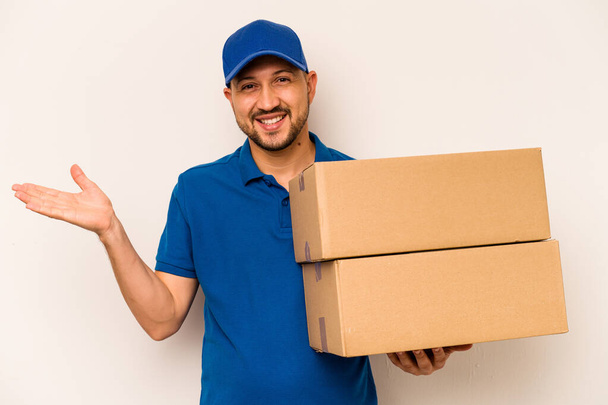 Hispanic delivery man isolated on white background showing a copy space on a palm and holding another hand on waist. - Photo, Image