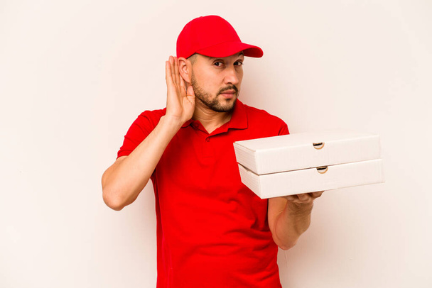 Young delivery man holding pizzas isolated on beige background trying to listening a gossip. - Photo, image