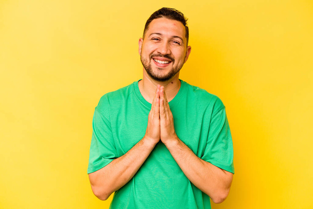 Young hispanic man isolated on yellow background holding hands in pray near mouth, feels confident. - Foto, immagini