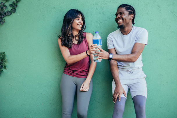 Two athletes resting after hard street workout session leaning on a stone wall. African american man and indian woman resting and drinking water - Photo, Image