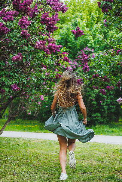 Egy gyönyörű göndör nő nyári ruhában fut és örül, napszemüveget visel. Egy lila lila lila bokor hátterében - Fotó, kép