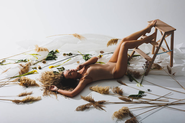 A slender woman in a beige jumpsuit poses next to dried flowers on a white isolated background. Sportswoman in a beige swimsuit posing next to flowers on a white background. - Foto, Imagem