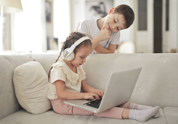 little girl works with a computer sitting on the sofa while her brother watches - Φωτογραφία, εικόνα