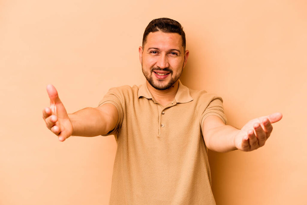 Joven hombre hispano aislado sobre fondo beige mostrando una expresión de bienvenida. - Foto, imagen
