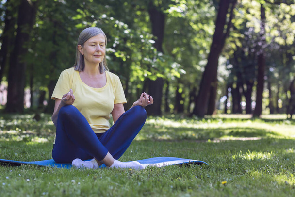 Időskorú, ősz hajú, nyugdíjas nő a parkban meditál és légzési gyakorlatokat végez, nyári lótuszpozícióban. - Fotó, kép