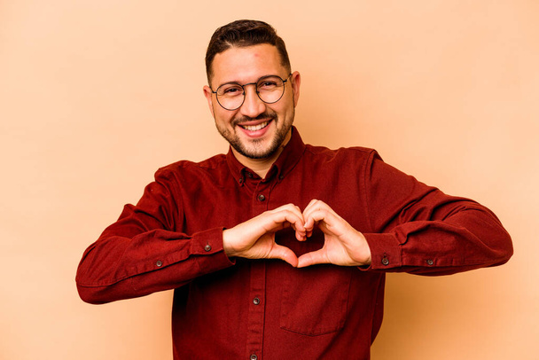Jonge Spaanse man geïsoleerd op beige achtergrond glimlachend en toont een hartvorm met handen. - Foto, afbeelding