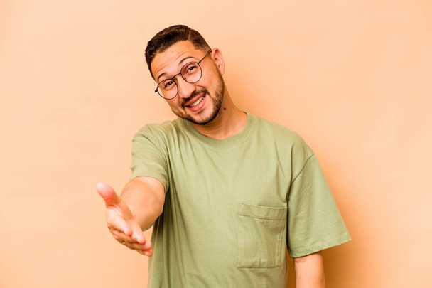 jonge latino man geïsoleerd op beige achtergrond stretching hand op camera in groet gebaar. - Foto, afbeelding