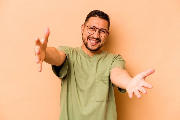Young hispanic man isolated on beige background feels confident giving a hug to the camera. - Fotografie, Obrázek