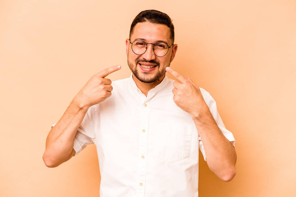 Young hispanic man isolated on beige background smiles, pointing fingers at mouth. - Fotó, kép