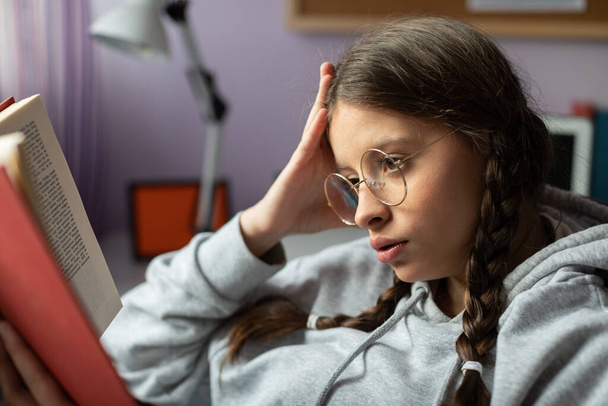A teenage girl with glasses reads her next school reading with interest. - Zdjęcie, obraz