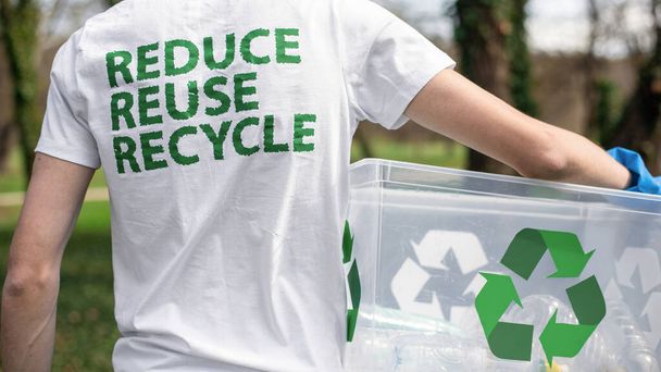 Hombre en la recolección de basura de plástico en un parque contaminado. Guantes de goma, sosteniendo un recipiente con botellas de plástico. Carteles de reciclaje en la camiseta y el contenedor - Foto, imagen