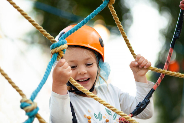 Little girl in helmet climbs ropes in adventure park outdoors. Extreme sport, active leisure on nature. - Foto, afbeelding