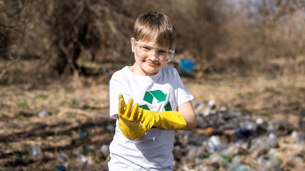 Poika turvalaseissa laittaa kumihanskat muovijätettä keräämiseen saastuneessa raivauksessa, kierrätyskyltti t-paidassa - Valokuva, kuva