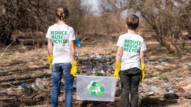 Ragazzo e ragazza in possesso di un contenitore presso la raccolta dei rifiuti di plastica in una radura inquinata, segni di riciclaggio sulle T-shirt - Foto, immagini